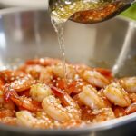 Sesame-garlic marinade being poured over cooked shrimp in a metal bowl