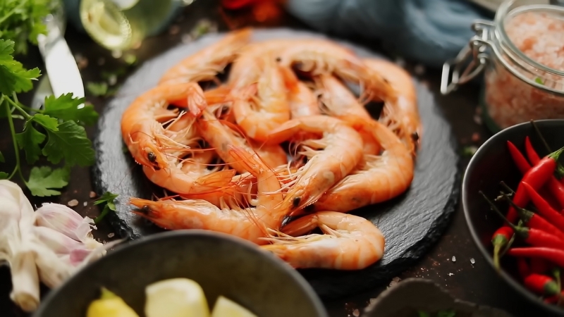 Fresh jumbo shrimp arranged on a dark slate plate, surrounded by garlic, chili peppers, lemon, and herbs