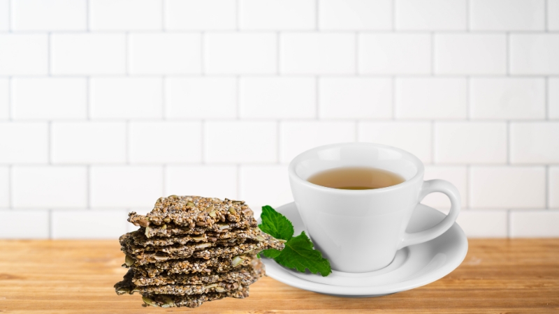 A stack of chia seed crackers sits next to a cup of unsweetened green tea on a wooden surface with a white tile background