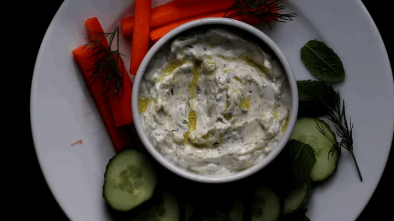 A bowl of chilled Greek yogurt dip with dill served on a plate with carrot sticks, cucumber slices, and fresh herbs