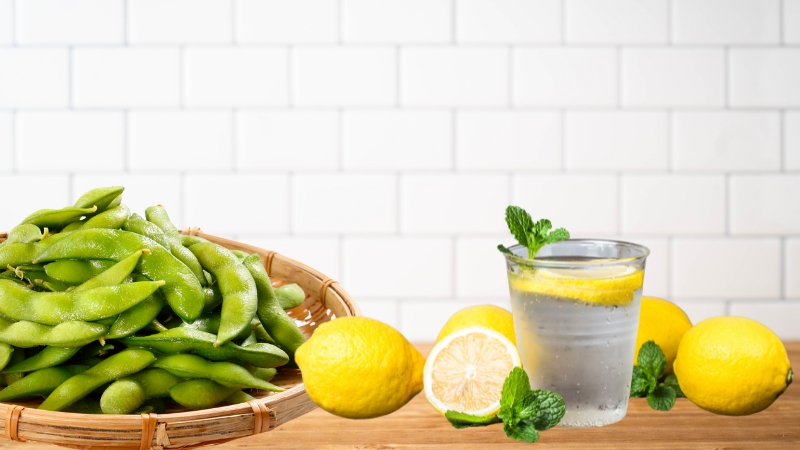 A basket of roasted edamame with sea salt sits next to a glass of citrus-infused water with lemons and mint on a wooden surface