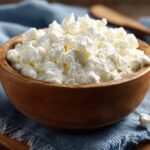 A wooden bowl filled with cottage cheese placed on a blue cloth napkin with a wooden spoon in the background