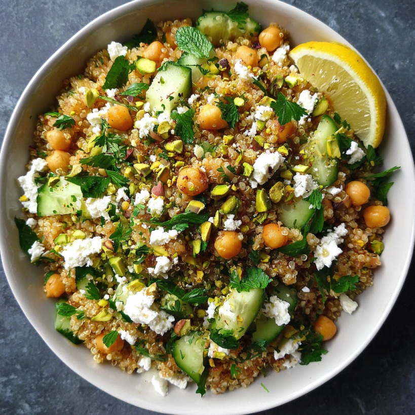 Colorful quinoa salad with chickpeas, cucumbers, feta, mint, pistachios, and lemon wedge in a white bowl